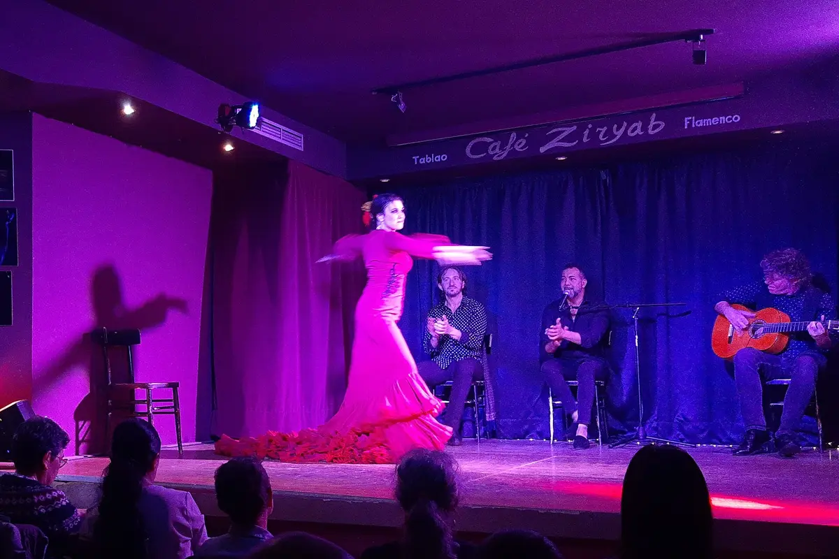 A dancer in a long pink dress with ruffled train spinning on a tablao stage, with a clapping singer and a guitarist seated behind her and audience heads in front