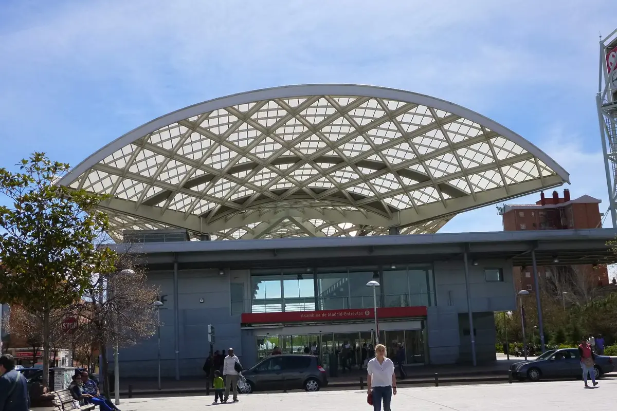 The glass-and-steel arched roof of the Asamblea de Madrid-Entrevías station building, with a red station sign above its entrance and a paved square in front