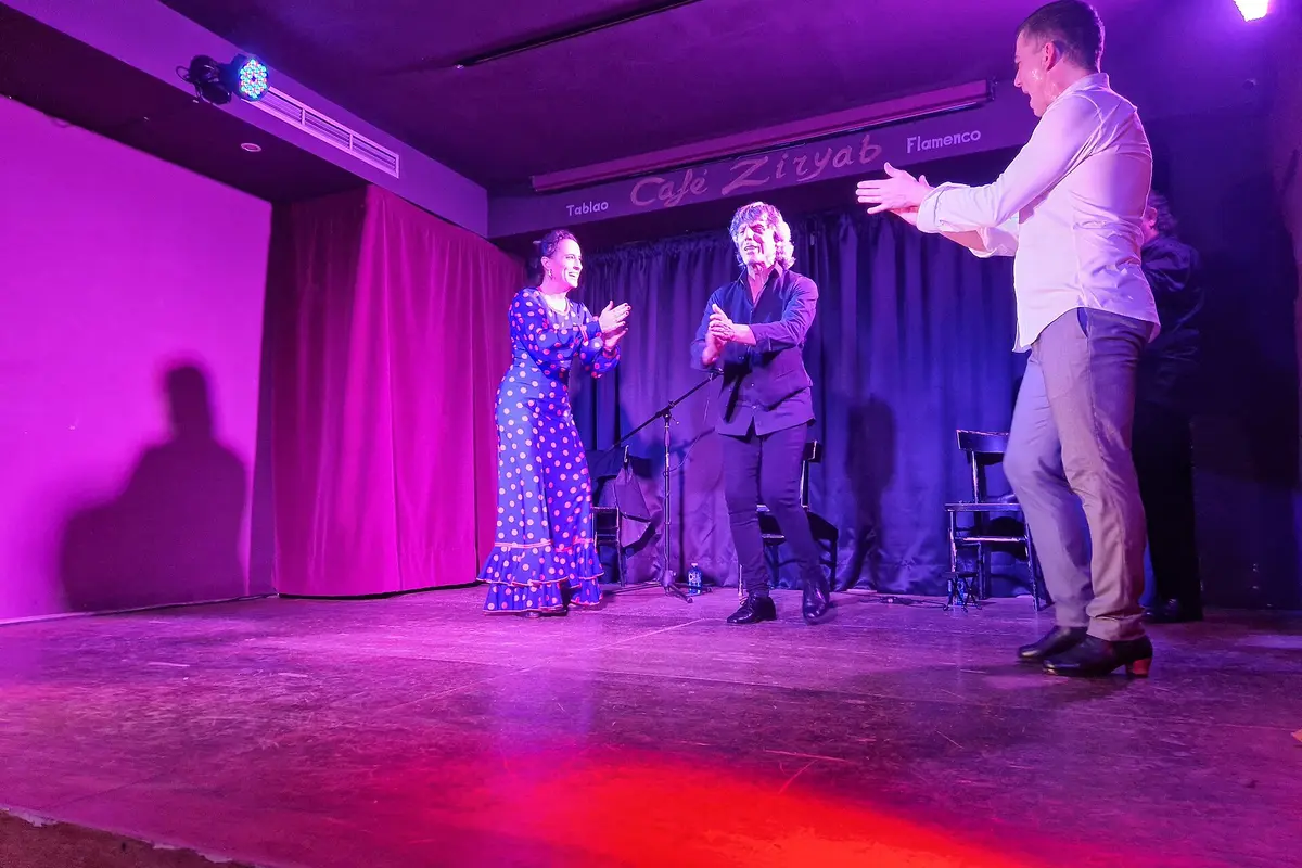 Two dancers and a singer clapping on a small tablao stage lit in purple and red, with a dark blue curtain behind them